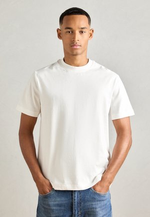 Jeune homme aux cheveux courts, debout avec les mains dans les poches de son jean, portant un t-shirt blanc à manches courtes contre un fond clair.