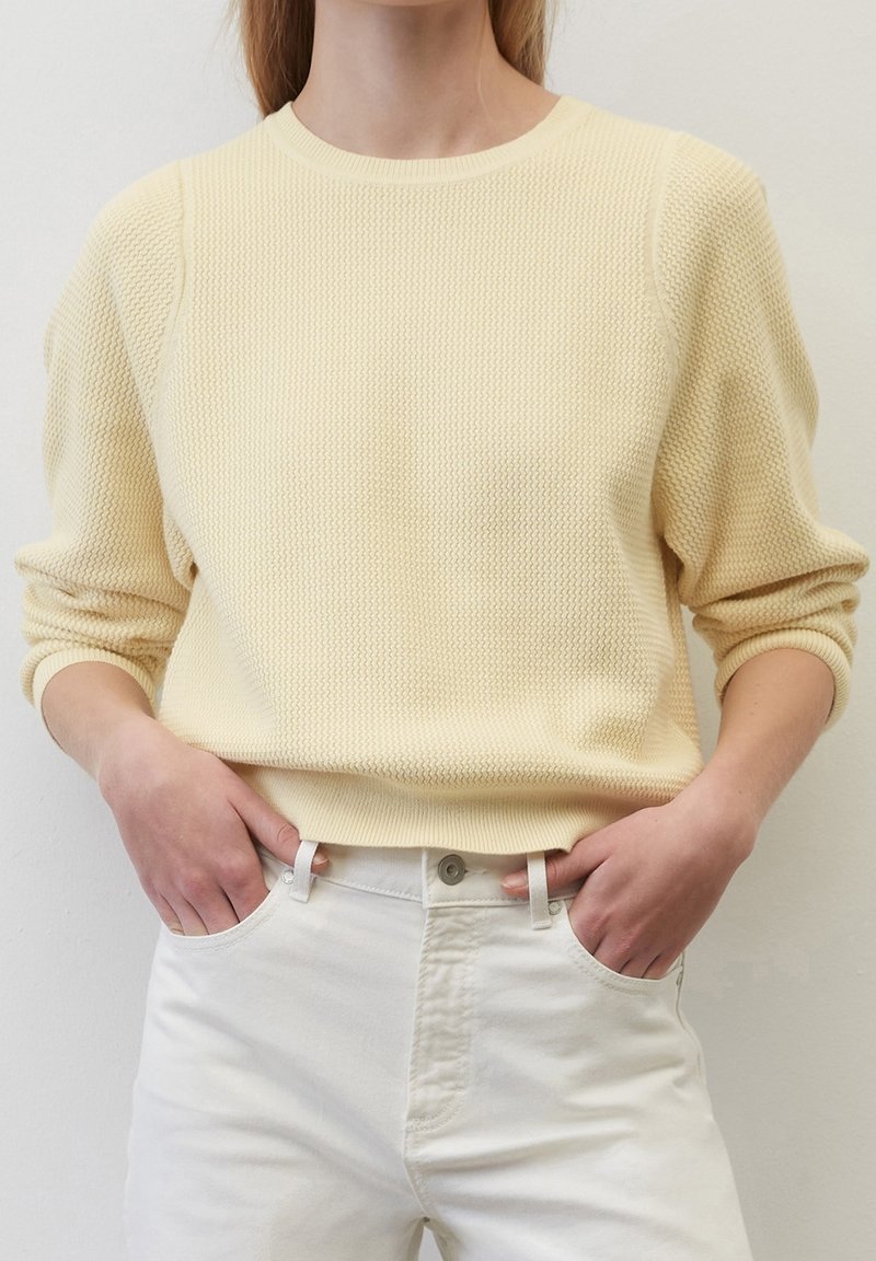 Personne portant un pull à manches longues texturé jaune pâle et un pantalon blanc taille haute, les mains dans les poches.