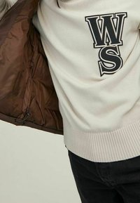 Beige tröja med stora svarta "WS" bokstäver, bärs under en brun jacka och kombineras med svarta byxor.