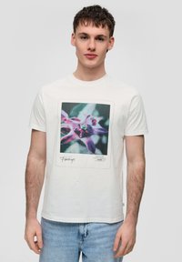 Witte katoenen T-shirt met een kleurrijke bloemenafdruk in het midden van de borst, korte mouwen en een ronde halslijn. Blauwe jeans gedragen.