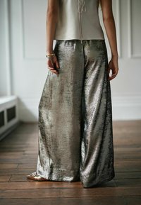 Metallilised laiad püksid, mis on läikiva tekstuuriga hõbedased, lõdva lõike ja sügavate volditega, koos heledat tooni pealsetega.
