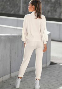 Cremefarbenes Loungewear-Set mit einem Pullover mit hohem Kragen und schlank geschnittenen Jogginghosen, mit gerippten Bündchen und einer Rückentasche. Weiße Knöchelstiefel.