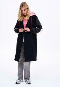 Mladá žena v dlouhém černém kabátu s nadýchanými rukávy, růžové mikině s kapucí, růžovém svetru, šedých džínách a stříbrných teniskách stojí před bílým pozadím.