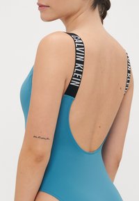 Mėlynas vientisas maudymosi kostiumėlis su gilia nugara ir juodomis dirželių juostomis, ant kurių yra balta "CALVIN KLEIN" prekės ženklo etiketė. Lygus paviršius ir prigludusi forma.