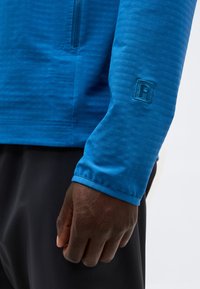 Κοντινό πλάνο ενός σκουρόχρωμου χεριού που φοράει μπλε μπλούζα με μακριά μανίκια, φερμουάρ και κεντημένο γράμμα "R" στο μανίκι.