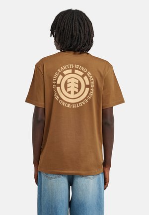 Personne à la peau foncée et aux dreadlocks porte un t-shirt marron avec un graphisme circulaire et les mots "FIRE EARTH WIND WATER" au dos.