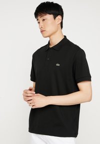 Čierna polo tričko vyrobené z bavlny, s trojitém gombíkom, krátkymi rukávmi a malým zeleným logom na ľavej hrudi.