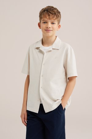 Chłopiec z krótkimi kręconymi włosami, ubrany w jasnobeżową, krótką koszulę na guziki, noszoną na białej koszulce i ciemnoniebieskich spodniach, uśmiecha się z jedną ręką w kieszeni.