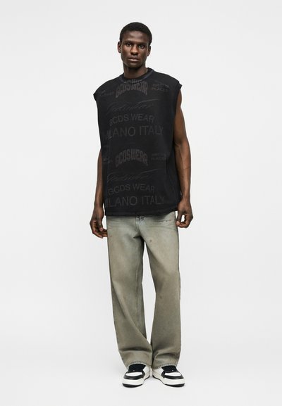 Άνδρας που φοράει μαύρο αμάνικο τυπωμένο πουκάμισο, φαρδιά ξεθωριασμένα γκρι τζιν και μαύρα-άσπρα αθλητικά παπούτσια, στέκεται μπροστά σε απλό φόντο.