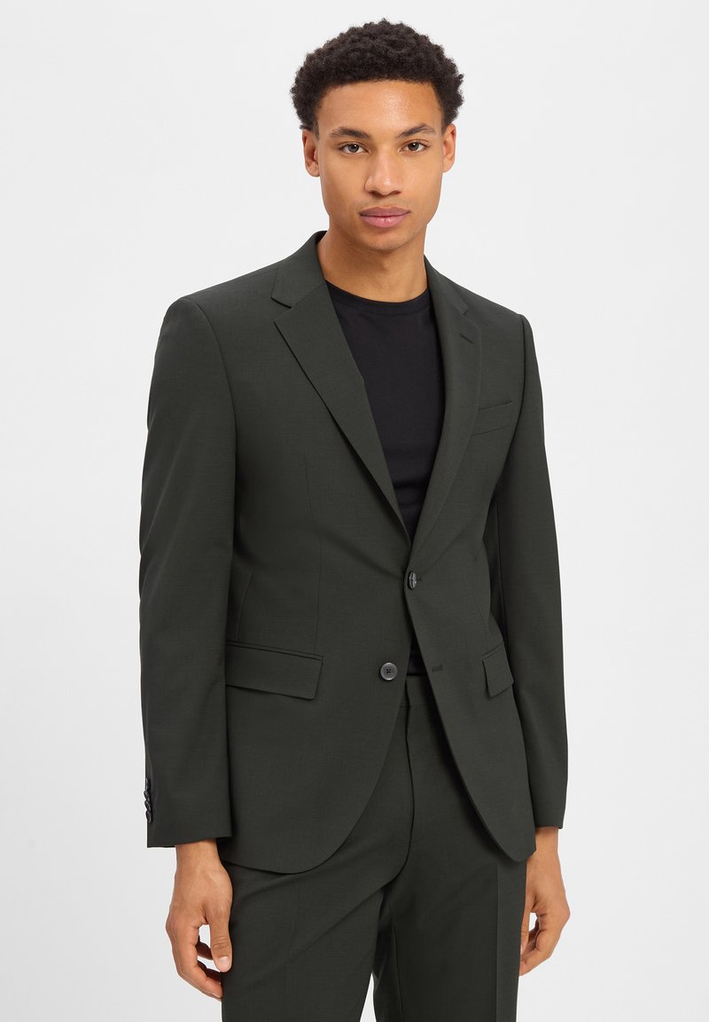 Ciemnozielony garnitur na miarę z wyciętymi klapami, zapinany na jeden guzik, z dwoma kieszeniami i dopasowanym krojem. Nałożony na czarną koszulę.