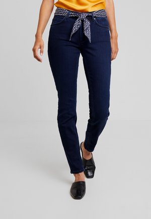 Kvinde iført mørkeblå skinny jeans med mønstret stofbælte, orange top og sorte teksturerede slip-on sko går fremad.