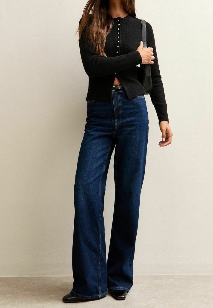 Femme portant un cardigan noir à manches longues boutonné, un jean bleu foncé à taille haute et jambes larges, des chaussures noires pointues, et tenant un petit sac à main noir.