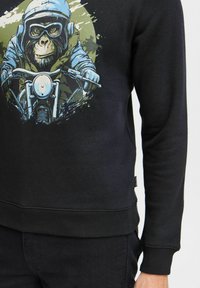 Felpa nera con una grafica di una scimmia in un casco blu, che guida una motocicletta, con dettagli in sfumature di verde e blu.