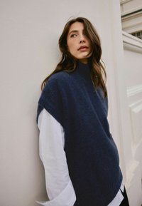 Junge Frau mit langen braunen Haaren, die einen marineblauen ärmellosen Pullover über einem weißen Langarmshirt trägt und sich an eine hellfarbige Wand lehnt.