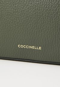 Borsa in pelle verde testurizzata con logo dorato "Coccinelle". Presenta una superficie liscia e resistente e una forma elegante.