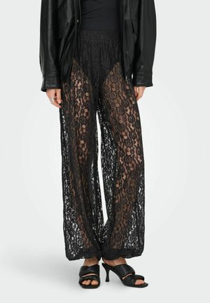 Femme portant un pantalon large en dentelle noire à motifs floraux par-dessus un short nude, un haut noir, une veste en cuir et des sandales à talons noires.