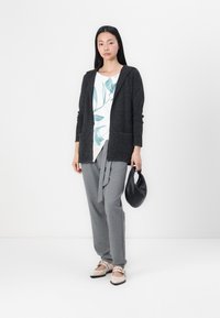 Dark grey, hooded cardigan with pockets, layered over white shirt featuring green leaf print. Paired with grey pants and beige flats.