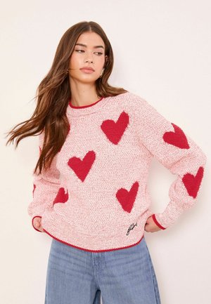 Frau trägt einen pinkfarbenen Strickpullover mit roten Herzen und rotem Saum, kombiniert mit blauer Jeans, steht mit den Händen in den Hüften.