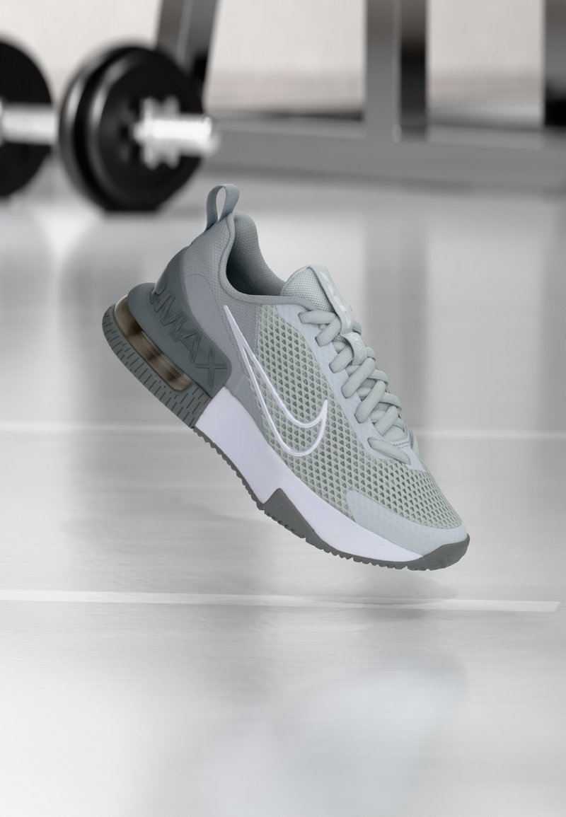 Baskets Nike Air Max gris clair avec tige en mesh et semelle à air visible, exposées dans une salle de sport avec des haltères floues à l'arrière-plan.