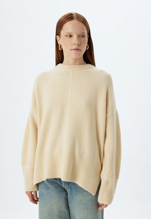 Beiger Strickpullover mit lockerer Passform, geripptem Saum und weißen Stichtdetails an den Schultern und am Rücken. Kombiniert mit hellblauen Jeans.