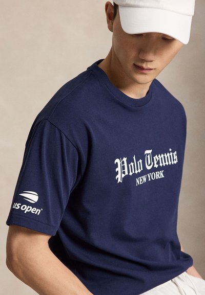 Νέος άνδρας με μπλε σκούρο μπλουζάκι Polo Tennis New York και λευκό καπέλο, κοιτάζει προς τα κάτω με ουδέτερο φόντο.