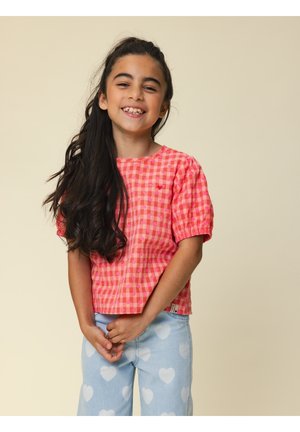 Lachend meisje met lang donker haar, gekleed in een rood geruit shirt en lichtblauwe broek met witte hartjes, staand tegen een beige achtergrond.