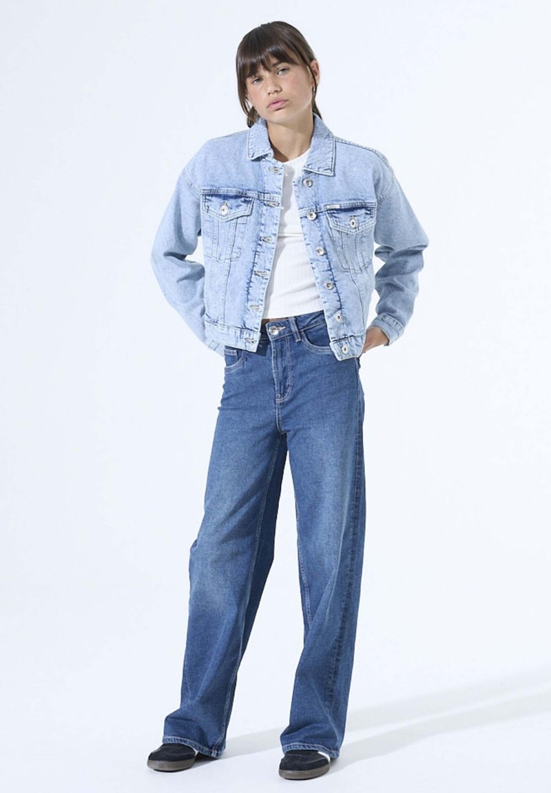 Light blue denim jacket with front pockets and buttons, paired with wide-leg blue denim jeans. Model wearing a white top and black shoes.