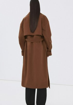 Mujer con cabello largo y liso, vistiendo un abrigo trench marrón con cinturón y pantalones negros, de pie de espaldas sobre un fondo liso.