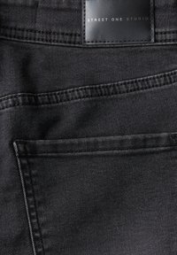 Zwarte denimstof met een gladde textuur, met een gestikte zak en een label met de tekst "STREET ONE STUDIO" aan de bovenkant.
