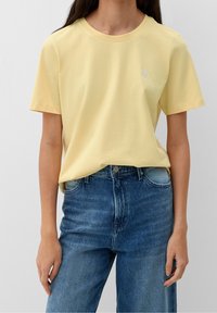Camiseta de algodón amarilla con manga corta, cuello redondo y un sutil gráfico de corazón en el lado izquierdo del pecho. Combinada con unos pantalones vaqueros de denim azul de talle alto.
