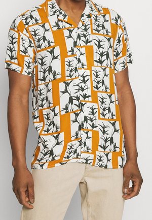 Homme portant une chemise à manches courtes avec un motif géométrique orange et blanc présentant des silhouettes de plantes noires et un pantalon beige clair.