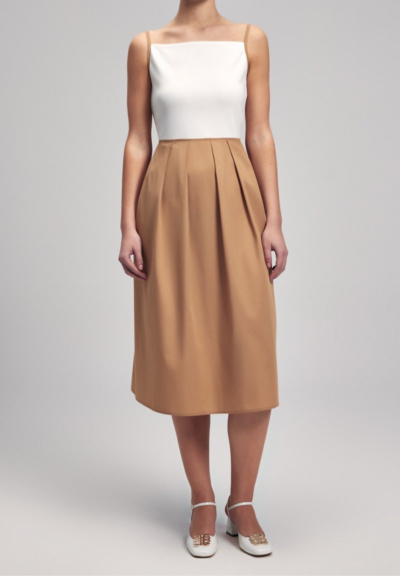 Femme portant un haut blanc sans manches rentré dans une jupe midi plissée couleur beige à taille haute, avec des chaussures blanches à talons carrés.