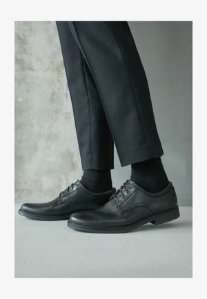 Schwarze Lederdressschuhe mit glatter Oberfläche, Schnürdesign, abgerundeter Zehenform und niedrigem Absatz, getragen mit schwarzen Anzughosen und Socken.