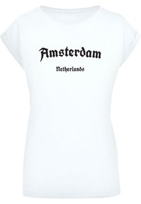 Valkoinen lyhythihainen t-paita, jossa on "Amsterdam" mustalla goottikirjaimella ja "Netherlands" pienemmässä mustassa tekstissä alla. Pehmeää puuvillamateriaalia.