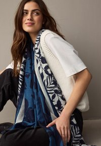 O eșarfă cu model alb și albastru, așezată peste un pulover alb din tricot, cu design floral și un material texturat. Pantaloni negri vizibili.