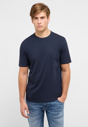 Nooruk helepruunide juustega, seljas tumesinine lühikeste varrukatega T-särk ja sinised teksad, seisab ühevärvilise heleda tausta ees.