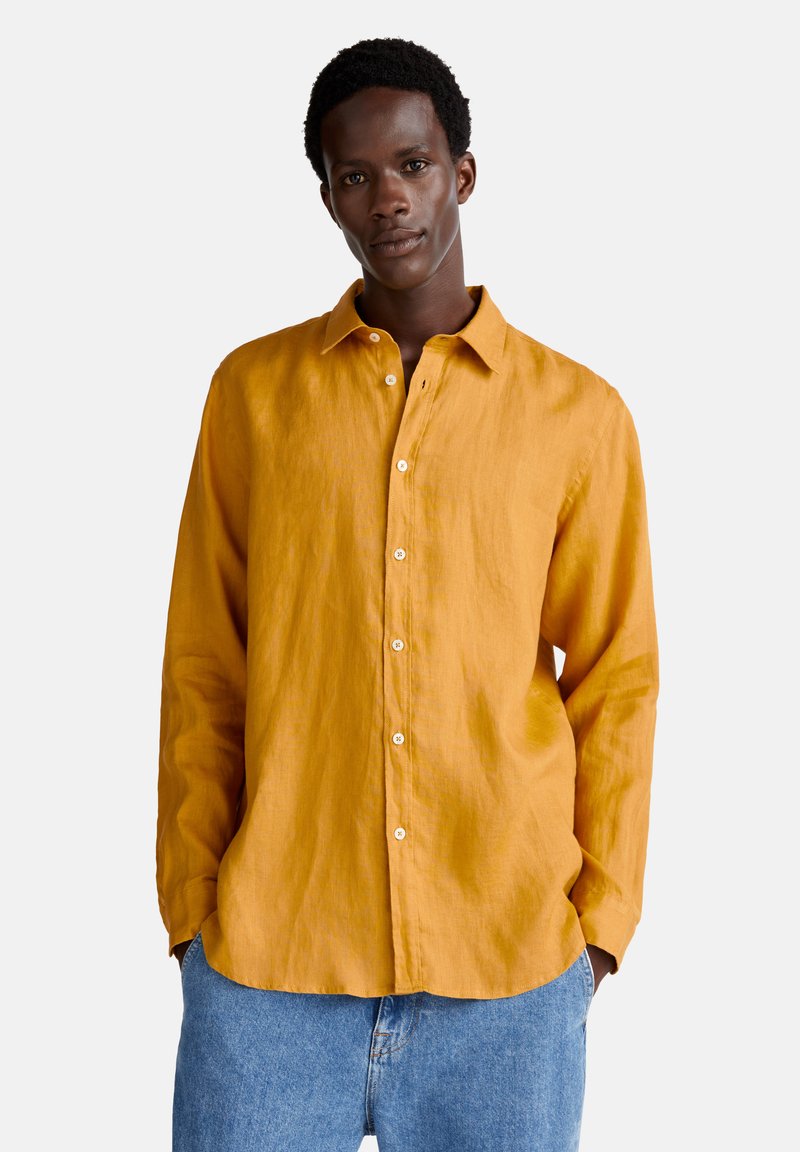 Camicia gialla in lino a maniche lunghe, vestibilità rilassata, con bottoni bianchi e colletto a punta, indossata sopra jeans di denim blu.