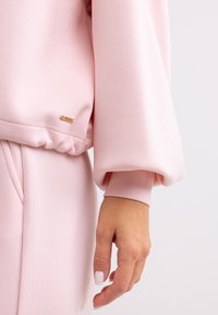 Sudadera rosa claro con un dobladillo fruncido y mangas anchas, que presenta una pequeña etiqueta dorada en el lado, textura suave y diseño minimalista.