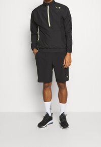 Svart lättviktsjacka med neongrön dragkedja och logotyp, tillsammans med svarta shorts. Svarta träningsskor och vita strumpor fulländar outfiten.