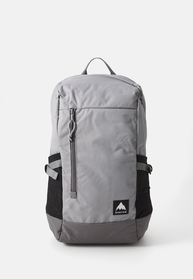 Sac à dos gris avec une poche avant zippée, des poches en mesh sur les côtés et une base noire. Présente un patch de logo et des bretelles ajustables.
