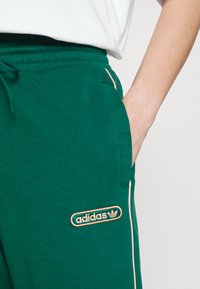 Pantalones de chándal verdes de algodón con una cinturilla elástica y cordón, que presentan un parche de logo con acento naranja y bolsillos laterales. Textura suave y corte relajado.