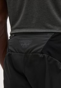 Κοντινό πλάνο ενός ατόμου που φοράει μαύρο αθλητικό σορτς με τσέπη με φερμουάρ και διακριτικό λογότυπο πάνθηρα στη μέση.