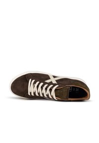 Sneaker in suede marrone con lacci bianchi e dettagli testurizzati. Presenta un logo in rilievo e una toppa sul tallone verde a contrasto. Suola in gomma piatta.