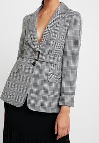 Svart och vit rutig kavaj med djup V-ringning, bälte i midjan, två framfickor och skräddarsydd design. Lättviktsmaterial med strukturerade axlar.