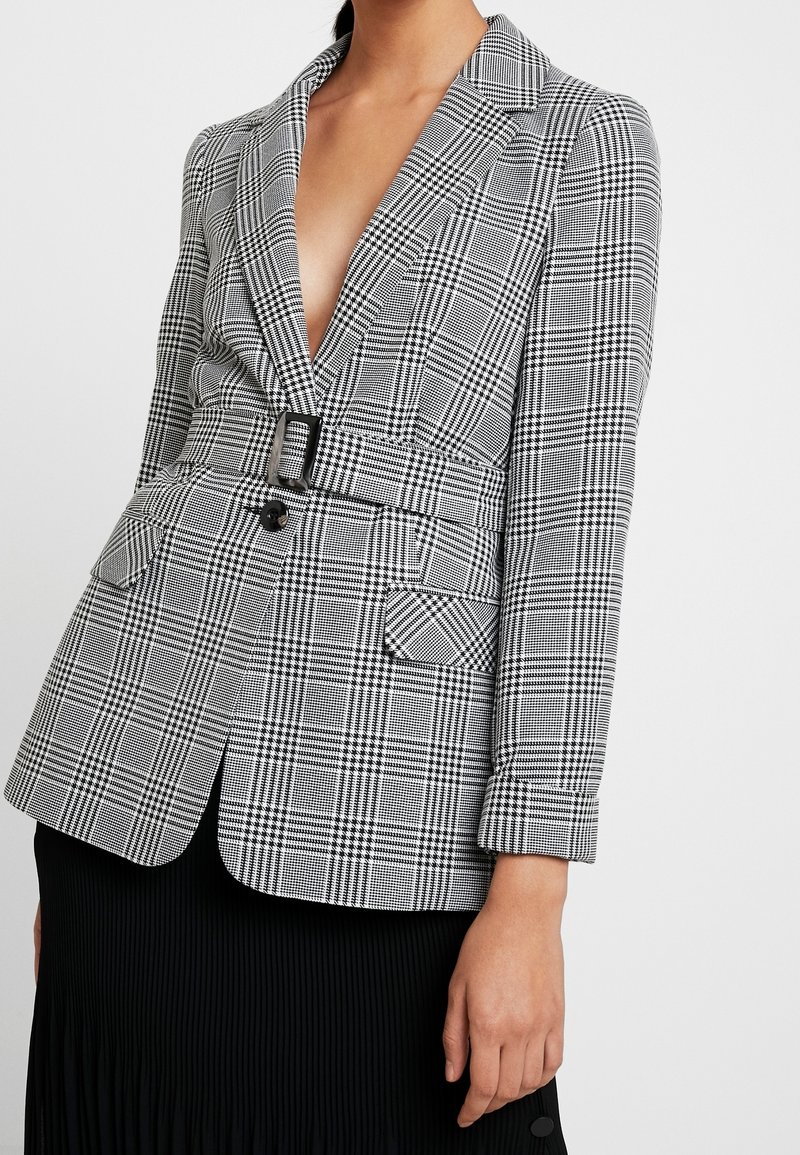 Svart och vit rutig kavaj med djup V-ringning, bälte i midjan, två framfickor och skräddarsydd design. Lättviktsmaterial med strukturerade axlar.