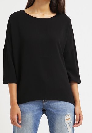 Kobieta ubrana w luźną czarną bluzkę z rękawem do łokcia, w połączeniu z jasnoniebieskimi postrzępionymi jeansami, na białym tle.