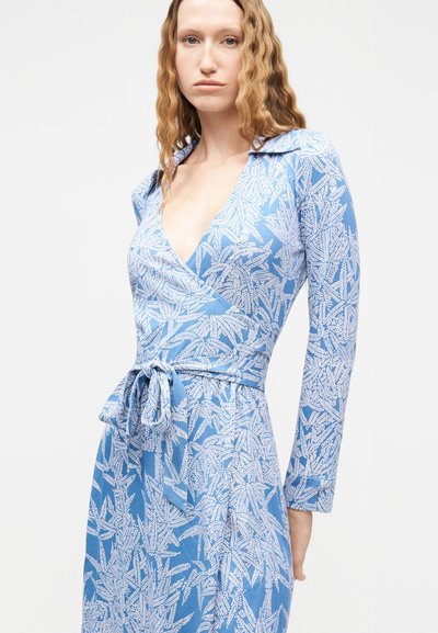 Γυναίκα με μακριά κυματιστά μαλλιά, φοράει μπλε φόρεμα τύπου wrap με λευκά φύλλα και ζώνη στη μέση, μπροστά σε απλό φόντο.