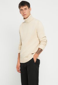 Beige genser med høy hals og ribbestrikkede mansjetter, med en teksturert strikk og sidesplitter, kombinert med svarte smale bukser.