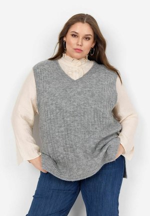 Chaleco de punto acanalado gris con diseño de escote en V, superpuesto a una blusa de gasa beige claro con volantes. El conjunto incluye unos pantalones vaqueros azules de corte relajado.