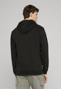 Crna majica s kapuljačom meke teksture, rebrastim manšetama i ravnim rubom. Vidljivi detalji šavova na vratu i ramenima.